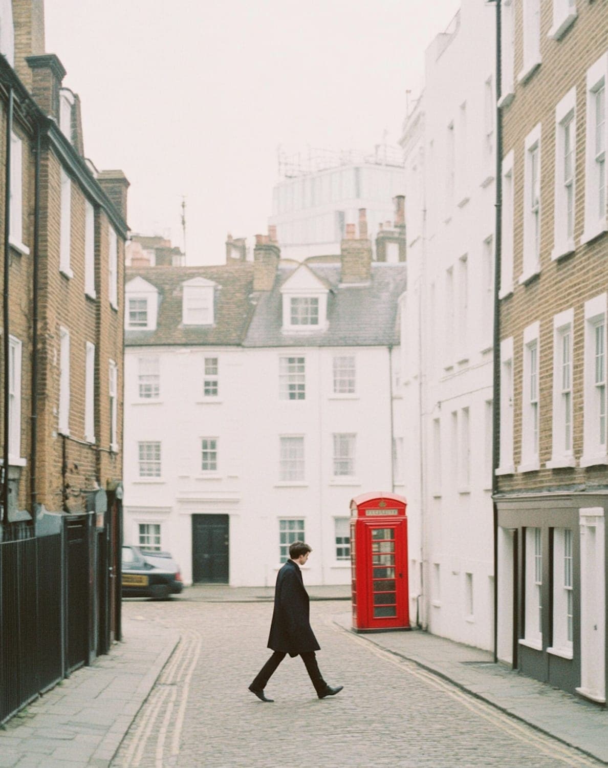 A man in a dark coat walking a cobbled London street past a red telephone box and pale terraced buildings